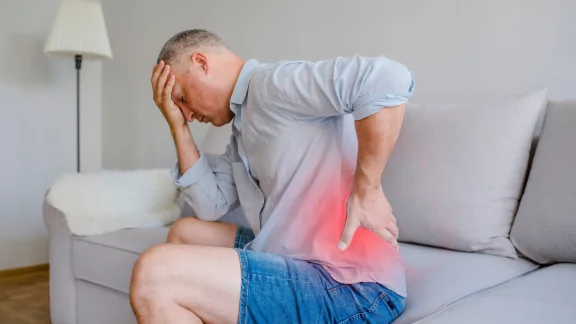 Man sitting on a couch holding his lower back in pain, area highlighted